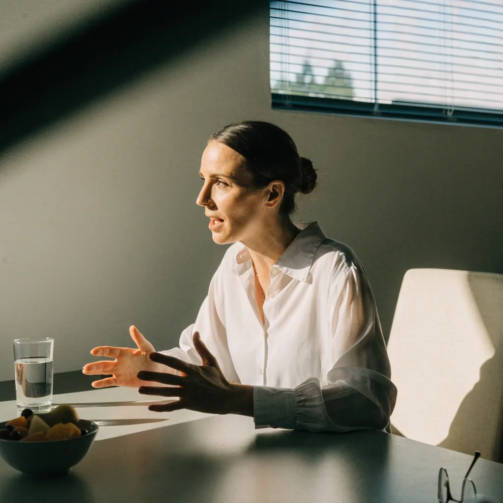 An empty consultation chair and a small table with a glass of water in a plain consulting room lit by hard midday light