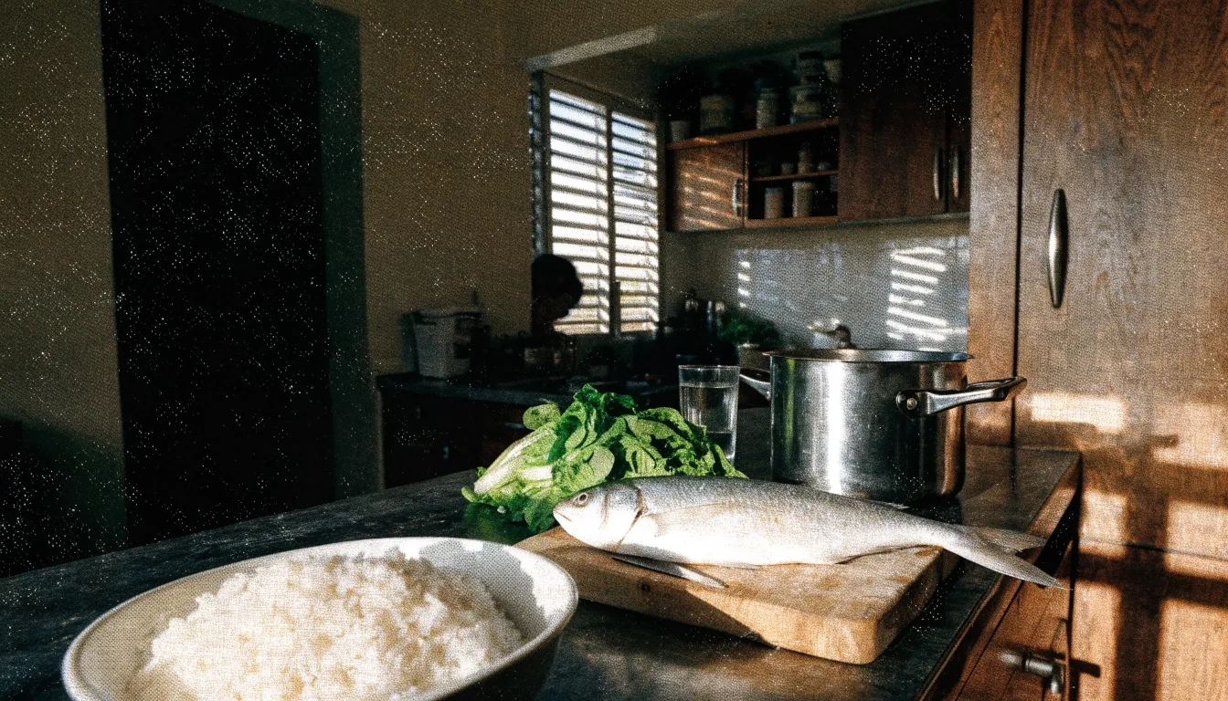 A home kitchen counter at noon with rice, a whole fish, cut greens, a steel pot and a glass of water